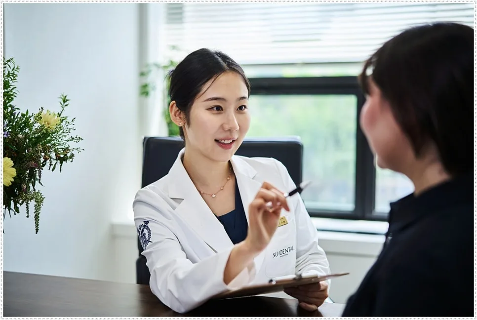 A dentist in a white coat holds a pen and clipboard as she smiles and talks with a patient across a desk.
