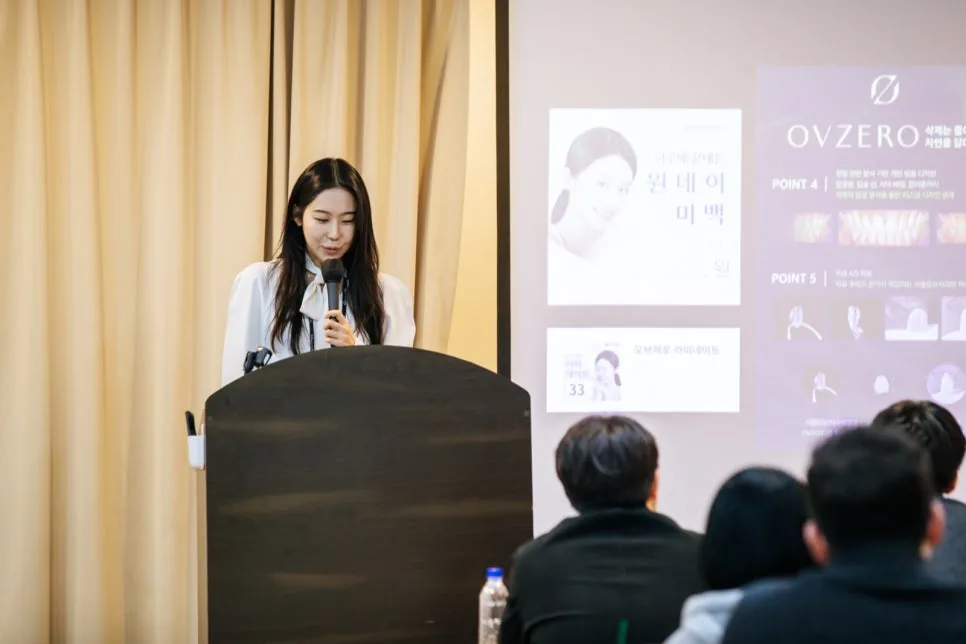 A dentist speaks into a microphone at a podium before an audience, with slides about one-day whitening and OVZERO projected behind her.
