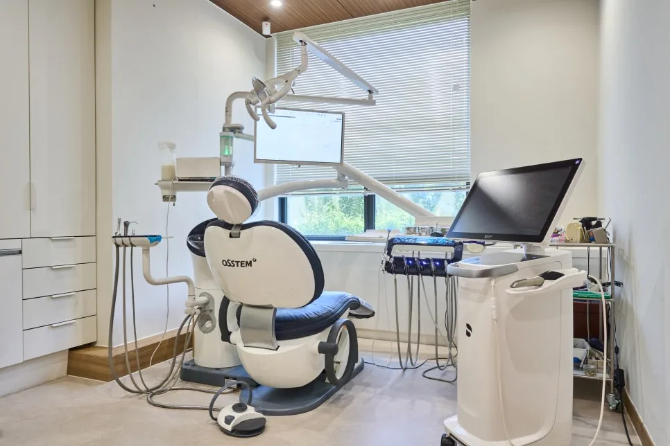 A bright treatment room with an Osstem dental chair, monitors and an intraoral scanner cart beside a window with blinds.