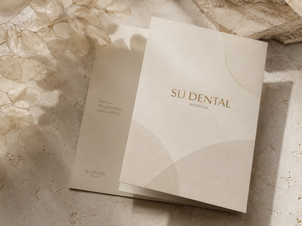 A cream-colored folded brochure with gold “SU DENTAL HOSPITAL” lettering rests on textured stone beside dried botanicals.
