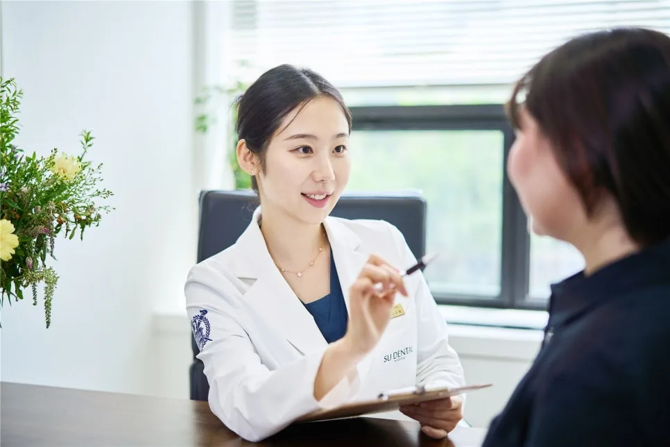 흰 가운을 입은 치과의사가 꽃이 놓인 창가 책상에서 펜과 클립보드를 들고 미소 지으며 환자와 상담하는 모습입니다.